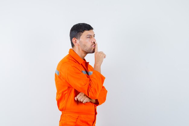 Trabajador joven mostrando gesto de silencio en uniforme y mirando con cuidado.