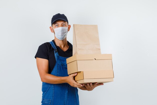 Trabajador joven sosteniendo cajas de cartón y bolsa de papel en uniforme, máscara, vista frontal.