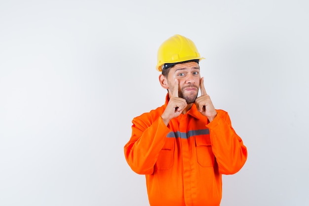 Trabajador joven en uniforme apuntando a sus párpados y mirando molesto.