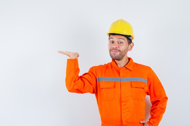 Trabajador joven en uniforme levantando la palma como sosteniendo o mostrando algo y mirando contento.