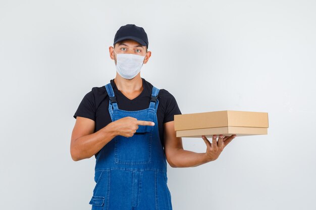 Trabajador joven en uniforme, máscara apuntando a caja de cartón, vista frontal.