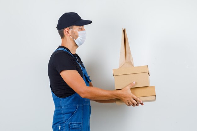 Trabajador joven en uniforme, máscara sosteniendo cajas de cartón y bolsa de papel y mirando serio.