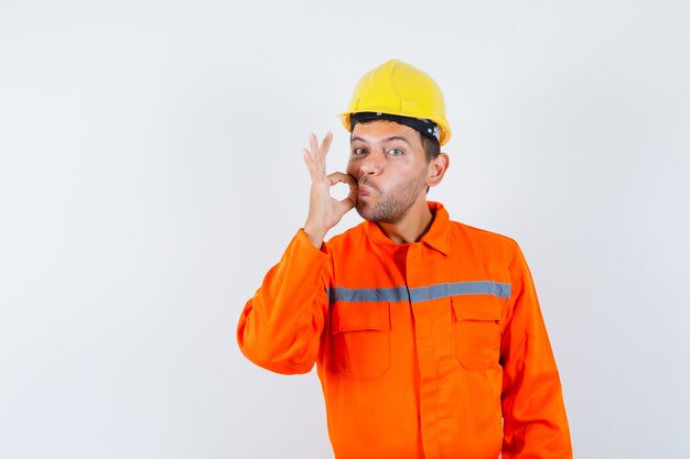 Trabajador joven en uniforme mostrando gesto de cremallera y mirando con cuidado.