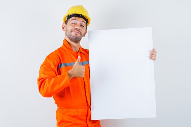 Trabajador joven en uniforme sosteniendo lienzo en blanco, mostrando el pulgar hacia arriba y luciendo alegre.