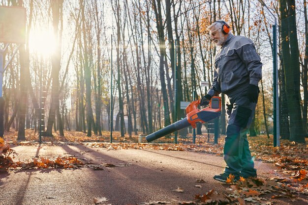 Trabajador de mantenimiento profesional en uniforme limpiando el parque de la ciudad de hojas secas de otoño con soplador de mano