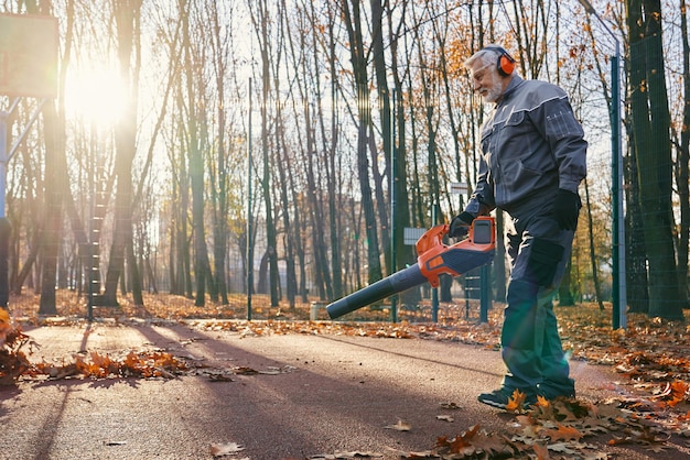 Trabajador de mantenimiento profesional en uniforme limpiando el parque de la ciudad de hojas secas de otoño con soplador de mano