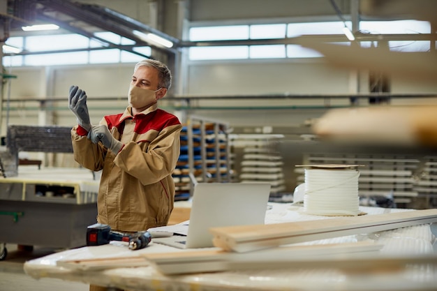 Trabajador manual poniéndose guantes protectores mientras trabaja en un taller de carpintería durante la pandemia del coronavirus