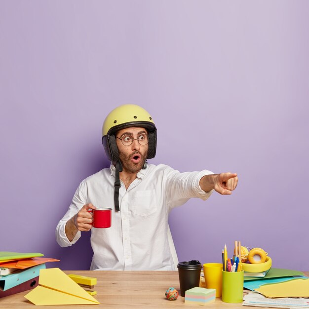 Trabajador masculino europeo emocional señala a la distancia, tiene expresión asustada, usa casco protector, sostiene una taza de café, trabaja en el escritorio de su gabinete