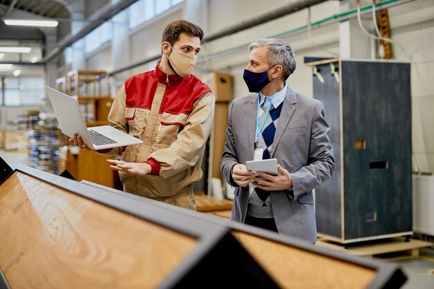Trabajador masculino que usa una computadora portátil mientras se comunica con el gerente de la empresa en la fábrica de madera durante la pandemia del coronavirus