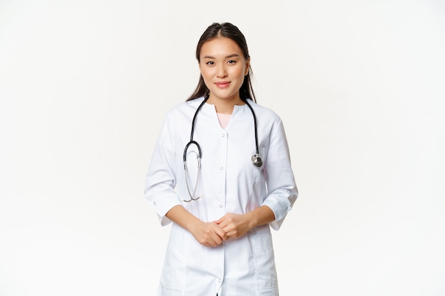 Trabajador médico asiático sonriente con estetoscopio, vestido con uniforme de médico, mirando útil al paciente, de pie sobre fondo blanco.