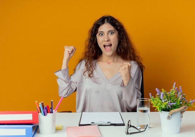 Trabajador de oficina bastante femenino joven feliz que se sienta en el escritorio con las herramientas de la oficina que muestra el gesto sí aislado en la pared naranja