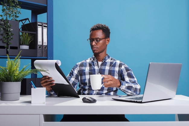 Trabajador de oficina de la empresa con portapapeles de documentación y taza de café mirando gráficos de gestión de proyectos. Joven empleado afroamericano sentado en un espacio de trabajo moderno mientras revisa el papeleo.
