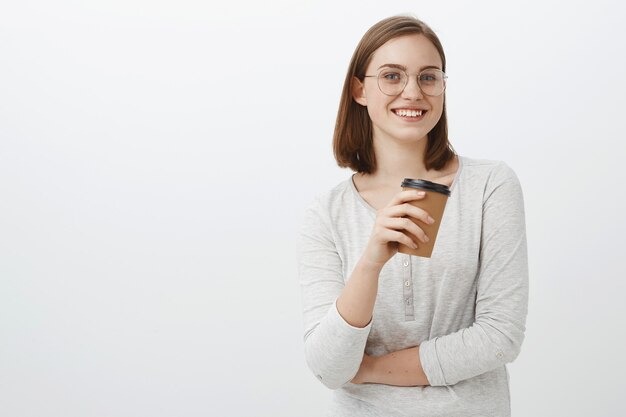 Trabajador de oficina femenino encantador feliz y entusiasta creativo que tiene descanso de pie en el café sobre la pared gris sosteniendo una taza de café de papel hablando divertido con un compañero de trabajo sonriendo