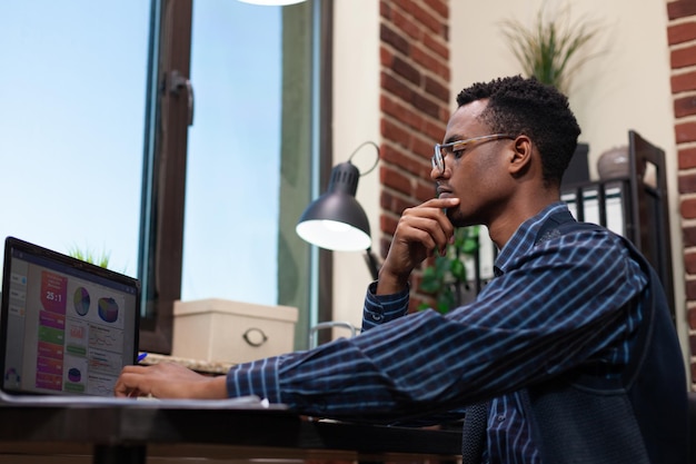 Trabajador de oficina de inicio afroamericano mirando centrado en la pantalla del portátil con analítica de negocios sentado en el escritorio. Especialista en marketing con la mano en la barbilla contemplando los indicadores clave de rendimiento.
