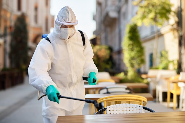 Trabajador de la salud con traje de materiales peligrosos que desinfecta un café en la acera durante la epidemia de COVID19