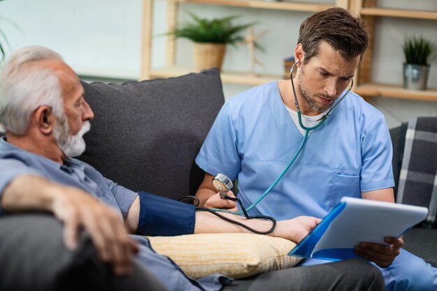 Trabajador sanitario serio que mide la presión arterial de un hombre maduro mientras revisa sus informes médicos durante una visita a su hogar