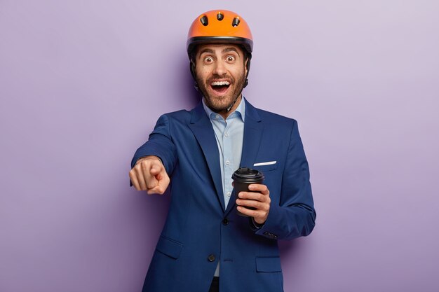 El trabajador técnico feliz apunta a la distancia, tiene una pausa para el café, usa traje y casco de construcción naranja, le indica que está en alto espíritu