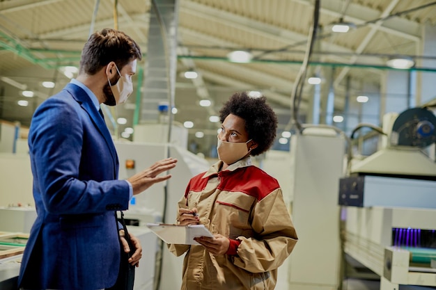 Trabajadora afroamericana y gerente de la empresa con máscaras faciales mientras habla en una fábrica de carpintería