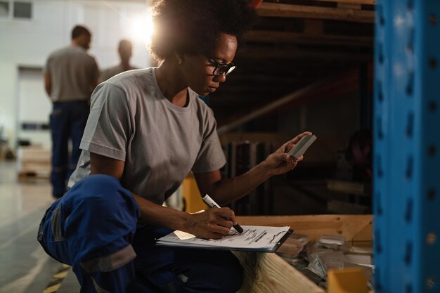 Trabajadora afroamericana revisando productos antes del envío y tomando notas en un almacén