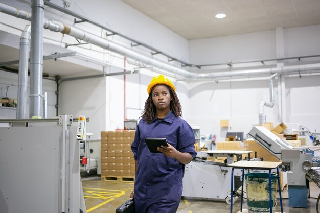 Trabajadora afroamericana seria en uniforme protector caminando al lugar de trabajo en el piso de la planta, sosteniendo la tableta y el estuche con herramientas