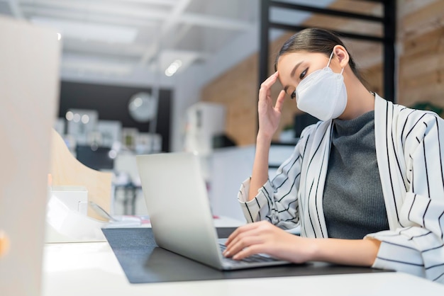 Trabajadora asiática con mascarilla que se siente infeliz, dolor de cabeza, dolor de cabeza, síndrome de oficina estresante e infrausto en la noche de la oficina por el concepto de trabajo