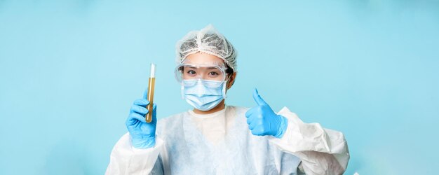 Trabajadora de laboratorio médica asiática en equipo de protección personal que muestra los pulgares hacia arriba y la muestra de prueba tu