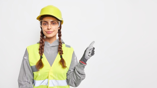 Trabajadora profesional ingeniera vestida con casco protector uniforme de trabajo, gafas transparentes y guantes indica en el espacio de la copia demuestra ideas para la construcción de edificios. Ingenieria