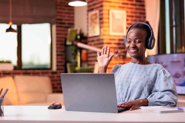 Una trabajadora remota afroamericana sonriente saluda a sus colegas en una videollamada en la oficina doméstica moderna. Reunión de videoconferencia con el equipo de la empresa, mujer de negocios con auriculares en teleconferencia