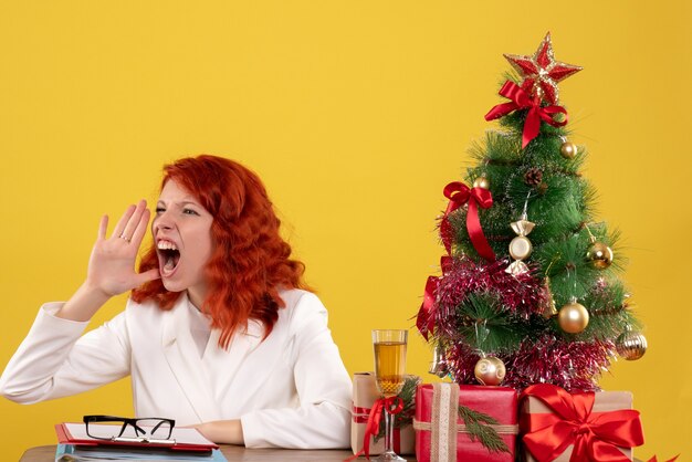 Trabajadora sentada detrás de la mesa con árbol de Navidad y presenta gritando en amarillo