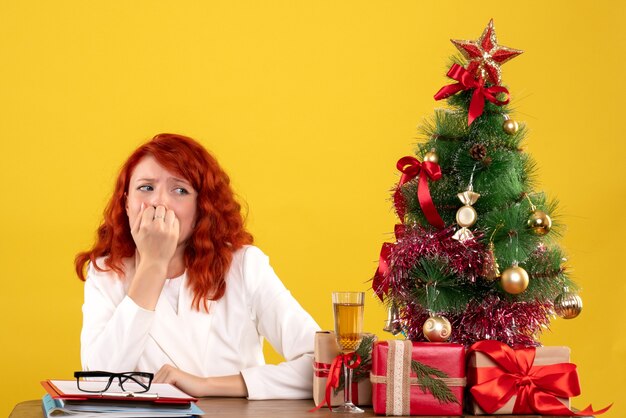 Trabajadora sentada detrás de la mesa con regalos de Navidad y árbol en amarillo