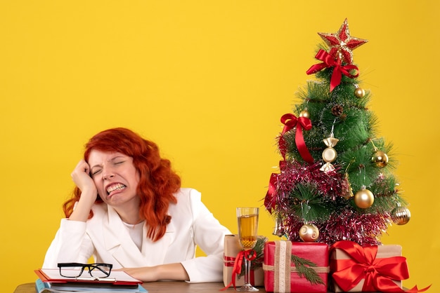 Trabajadora sentada detrás de la mesa con regalos de Navidad y árbol en amarillo