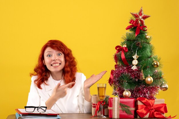 Trabajadora sentada detrás de la mesa con regalos de Navidad y árbol en amarillo