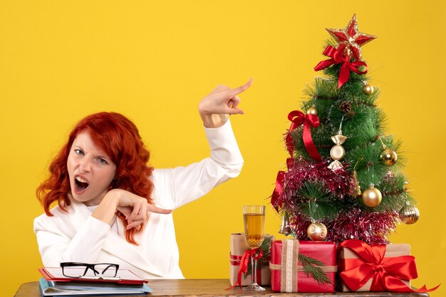 Trabajadora sentada detrás de la mesa con regalos de Navidad y árbol en amarillo