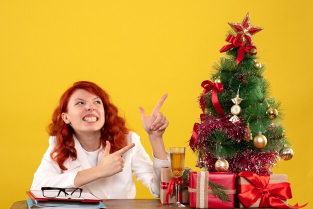 Trabajadora sentada detrás de la mesa con regalos de Navidad y árbol en amarillo