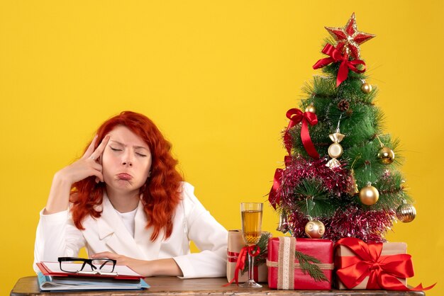 Trabajadora sentada detrás de la mesa con regalos de Navidad y árbol en amarillo
