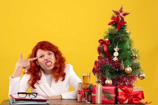 Trabajadora sentada detrás de la mesa con regalos de Navidad y árbol en amarillo