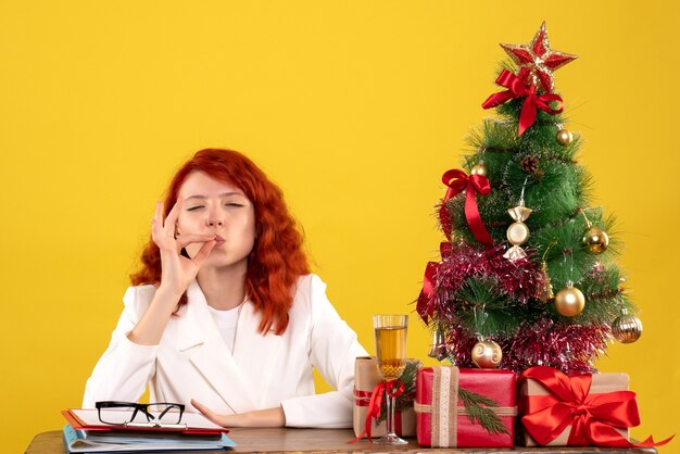 Trabajadora sentada detrás de la mesa con regalos de Navidad y árbol en amarillo