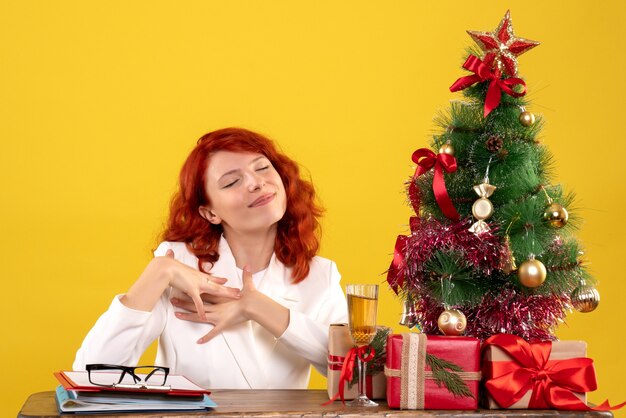 Trabajadora sentada detrás de la mesa con regalos de Navidad y árbol en amarillo