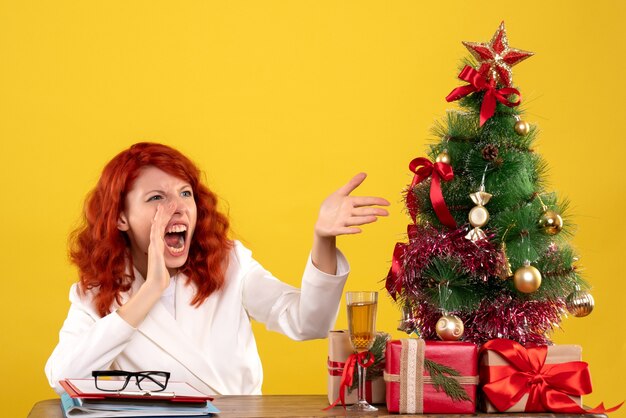 Trabajadora sentada detrás de la mesa con regalos de Navidad y árbol en amarillo