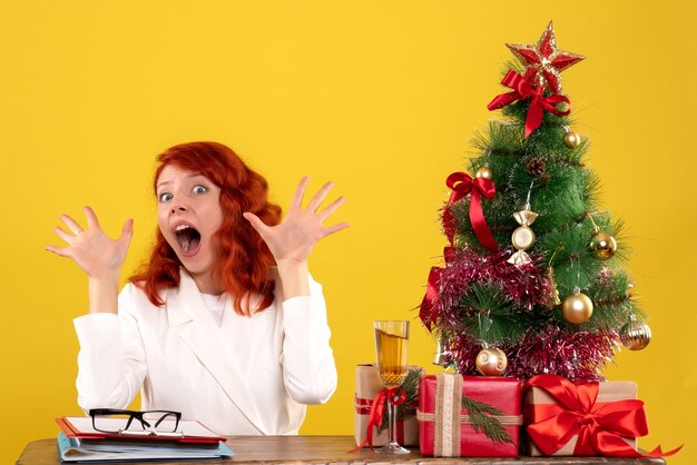 Trabajadora sentada detrás de la mesa con regalos de Navidad y árbol en amarillo