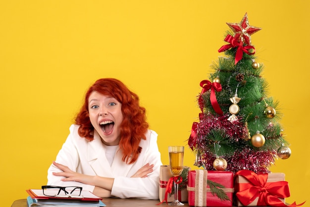 Trabajadora sentada detrás de la mesa con regalos de Navidad y árbol en amarillo