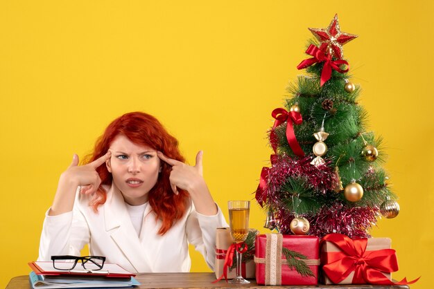 Trabajadora sentada detrás de la mesa con regalos de Navidad y árbol en amarillo