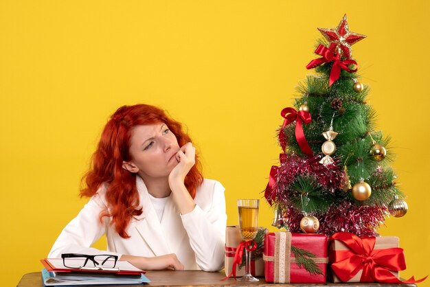 Trabajadora sentada detrás de la mesa con regalos de Navidad y árbol en amarillo