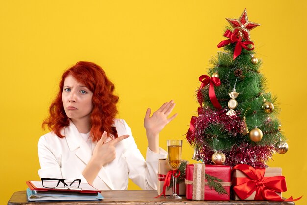 Trabajadora sentada detrás de la mesa con regalos de Navidad y árbol en amarillo