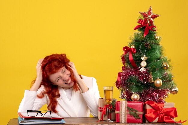 Trabajadora sentada detrás de la mesa con regalos de navidad y árbol en el piso amarillo oficina año nuevo color navidad