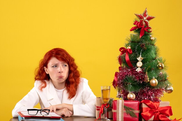 Trabajadora sentada detrás de la mesa con regalos de Navidad y árbol sintiéndose triste en amarillo