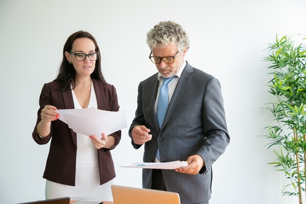 Trabajadores confiados en gafas leyendo documentos, hablando y de pie cerca de la mesa