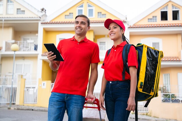 Trabajadores de correos sonrientes de pie y buscando la dirección de la casa en la tableta. Dos mensajeros felices entregando el pedido en bolsa térmica y vistiendo camisetas rojas. Servicio de entrega y concepto de compra online.