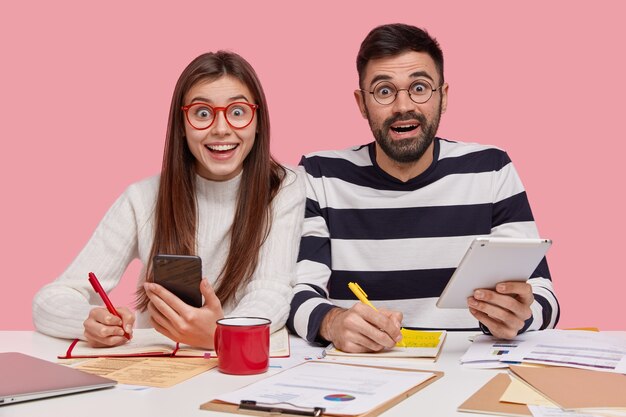 Los trabajadores del equipo se sientan en un escritorio, toman notas en el bloc de notas, se preparan para el seminario, sostienen un teléfono inteligente moderno y un panel táctil, tienen una mirada feliz, estudian documentos con gráficos, participan en el proceso de trabajo