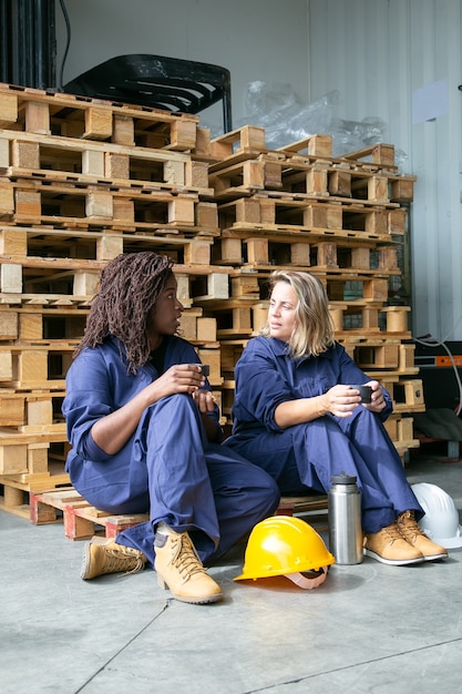 Foto gratuita los trabajadores de la fábrica charlando mientras beben café, comen galletas, sentados en un palet de madera en el almacén